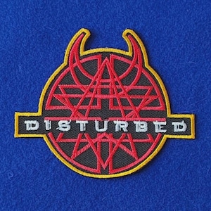 May include: A circular embroidered patch featuring a red and black geometric design with two upward-curving horns at the top. A horizontal black bar across the centre displays the white text DISTURBED, all outlined in a bright yellow border.