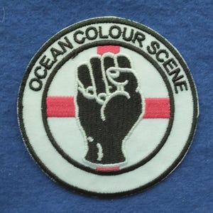 Puede incluir: Un parche bordado circular con fondo blanco y borde negro. El texto OCEAN COLOUR SCENE aparece arqueado en la parte superior. Un gráfico de un puño alzado negro está centrado sobre una franja horizontal roja en el medio.