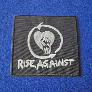 May include: A square black embroidered patch featuring a white circular arrow design surrounding a heart shape with a raised fist inside. The text RISE AGAINST is embroidered in white capital letters at the bottom of the patch.
