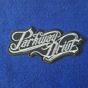 May include: An embroidered fabric patch featuring the text Parkway Drive in a stylised white script font with a black border. The lettering is interconnected and features decorative flourishes, creating a cohesive graphic design element.