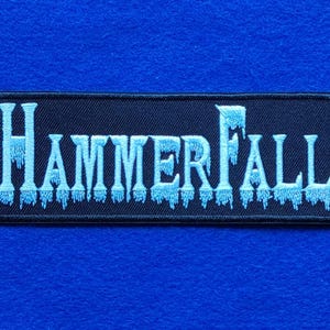 May include: A rectangular embroidered patch featuring the word HammerFall in light blue lettering. The text has a distinct icy, dripping effect at the bottom of each letter, set against a solid black background with a black stitched border.