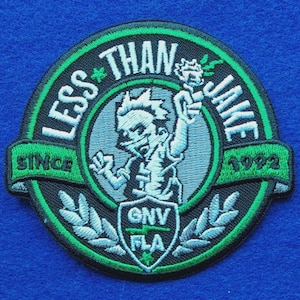 May include: A circular embroidered patch featuring a black and green colour scheme. The text reads Less Than Jake, Since 1992, and GNV FLA. A central figure holds a flaming object, surrounded by a border with laurel leaf designs and a small shield.