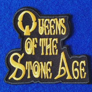 Peut inclure: Un écusson brodé noir avec le texte "QUEENS OF THE STONE AGE" en lettres dorées stylisées. L'inscription est cousue sur une base en tissu noir avec une bordure noire épaisse qui souligne la forme du lettrage.