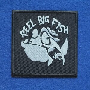 May include: A square black embroidered patch featuring a white graphic of a fish wearing a hat and glasses. The text REEL BIG FISH is arched above the fish illustration in white thread. The patch has a black stitched border on all four sides.