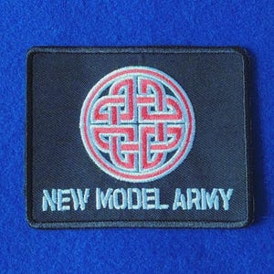 Op de afbeelding: Een rechthoekige zwarte geborduurde patch met een cirkelvormig rood en lichtblauw Keltisch knoopontwerp gecentreerd boven de tekst NEW MODEL ARMY in lichtblauwe hoofdletters. De patch heeft een zwarte gestikte rand en een plat oppervlak.