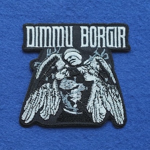Op de afbeelding: Een zwart-witte geborduurde patch met de tekst DIMMU BORGIR in een gestileerd lettertype aan de bovenkant. Onder de tekst staat een gedetailleerde afbeelding van een figuur met grote, gespreide gevederde vleugels, uitgevoerd in wit garen op zwart.
