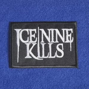 Op de afbeelding: Een rechthoekige zwarte stoffen patch met de wit geborduurde tekst ICE NINE KILLS in een gestileerd, druipend lettertype. De patch heeft een zwarte gestikte rand op een effen blauwe, getextureerde achtergrond.