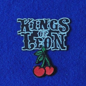 May include: An embroidered patch featuring the text Kings of Leon in black lettering with a light blue outline. Below the text is a graphic of two red cherries with green stems and leaves, all stitched with detailed thread work on a blue background.