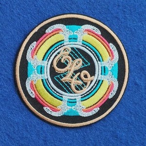Puede incluir: Un parche bordado circular con fondo negro y anillos concéntricos en turquesa, amarillo y rosa. El diseño incluye detalles de filigrana blanca y las letras cursivas color canela ELO centradas sobre un patrón de cuadrícula geométrica.