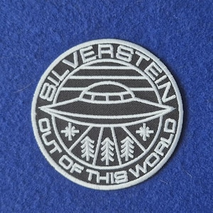 May include: A circular embroidered patch featuring a black background with white stitching. The design depicts a flying saucer above a forest of pine trees, with the text SILVERSTEIN and OUT OF THIS WORLD encircling the central graphic.
