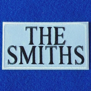 May include: A rectangular embroidered patch featuring a light blue background with a white border. The text THE SMITHS is stitched in black, bold, serif capital letters across the centre of the patch, creating a clear and legible design.