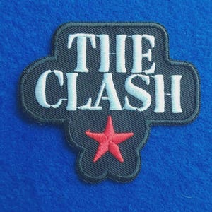 May include: A black embroidered patch with a shaped border featuring the white text THE CLASH in a bold serif font. Below the text is a single, five-pointed red star. The patch has a dark grey stitched edge and a textured fabric surface.