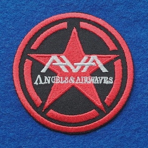 Op de afbeelding: Een ronde geborduurde patch met een rode ster op een zwarte achtergrond, omsloten door een rode cirkelvormige rand. De tekst AVA is in het wit geborduurd boven de tekst ANGELS & AIRWAVES, die ook in het wit over de rode ster is gestikt.