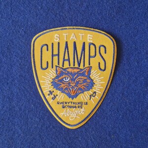 May include: A shield-shaped embroidered patch in yellow with a blue border. It features the text STATE CHAMPS, a central cat illustration, the letters NY and 70, and the text EVERYTHING IS GONNA BE Alright in blue and white thread.