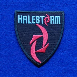 Puede incluir: Un parche bordado en forma de escudo con fondo negro y borde negro. El texto HALESTORM está bordado en azul claro, con un símbolo estilizado rojo que reemplaza la letra O. Un gran diseño gráfico rojo se encuentra debajo del texto.