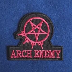 Puede incluir: Un parche bordado negro que presenta un símbolo de pentagrama rosa encerrado en un círculo con pequeñas púas que se extienden desde la parte inferior. El texto ARCH ENEMY está bordado en letras mayúsculas rosas en la base sobre fondo negro.