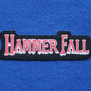 May include: A rectangular embroidered patch with a black background and the text "HAMMER FALL" stitched in red with a white outline. The lettering features a stylized font with a jagged, icy appearance along the bottom edges of the letters.