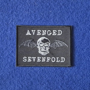 May include: A rectangular black embroidered patch featuring the white text AVENGED above a central white skull with bat wings, and the white text SEVENFOLD below the skull. The patch has a black stitched border and a textured fabric surface.