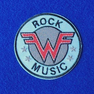 Op de afbeelding: Een ronde geborduurde patch met een lichtblauwe achtergrond en een zwarte rand. Het midden toont een grijze cirkel met een rood gestileerd W- en F-logo. De tekst ROCK staat in een boog erboven en MUSIC in een boog eronder in het zwart.