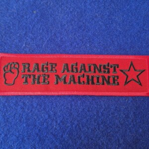 May include: A rectangular red embroidered patch featuring the black text RAGE AGAINST THE MACHINE. The design includes a black raised fist icon on the left side and a black five-pointed star outline on the right side, all on a red background.