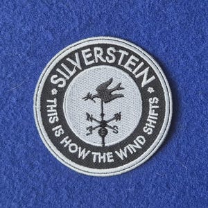 May include: A circular embroidered patch featuring a black and white weather vane design in the centre. The text SILVERSTEIN is arched at the top, and THIS IS HOW THE WIND SHIFTS is arched at the bottom, all set against a dark blue background.