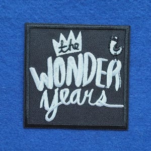May include: A square black embroidered patch featuring the white text "the WONDER years" in a stylised font. Above the text is a white crown icon, and to the right is a small white bird graphic. The patch has a black stitched border.