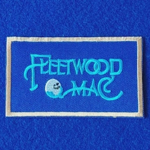 May include: A rectangular embroidered patch featuring a royal blue background with a light blue stitched border. The text Fleetwood Mac is embroidered in light blue, with a small white and light blue circular graphic integrated into the lettering.