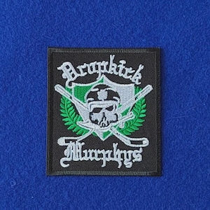 May include: A rectangular black embroidered patch featuring a white skull wearing a helmet, centred over a green laurel wreath and two crossed hockey sticks. The text Dropkick Murphys is stitched in white gothic lettering above and below.