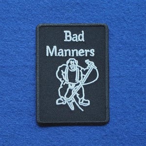 Op de afbeelding: Een rechthoekige zwarte geborduurde patch met de witte tekst "Bad Manners" boven een witte lijntekening van een figuur met bretels die een lang gereedschap vasthoudt, met een zwarte rand rondom het gehele ontwerp.