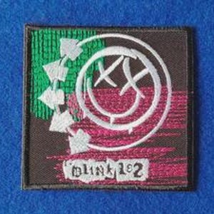 May include: A square embroidered patch featuring a black background with a white circular smiley face logo. The design includes a green textured area, pink horizontal stitching, and the text Blink-182 in a white rectangular banner at the bottom.