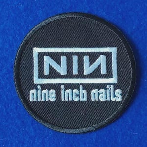 May include: A circular black embroidered patch featuring the white text NIN enclosed in a rectangular border, with the words nine inch nails printed in white lowercase letters directly underneath the box on a black fabric background.
