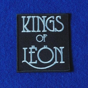May include: A square black embroidered patch featuring the white text KINGS OF LEON. The lettering is arranged in three lines with the word KINGS on top, OF in the middle, and LEON on the bottom, with a curved line underlining the word LEON.
