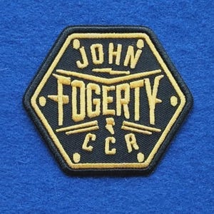 May include: A hexagonal embroidered patch featuring a black background with gold-coloured stitching. The text reads JOHN FOGERTY and CCR, accompanied by a graphic of two crossed drumsticks. The border is also stitched in a gold-coloured thread.