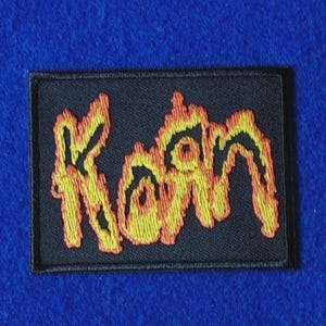 Puede incluir: Un parche rectangular bordado que presenta la palabra Korn en una fuente estilizada similar a llamas. Las letras están bordadas con hilo amarillo y naranja vibrante sobre un fondo negro sólido, con un borde negro cosido alrededor.