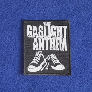 Op de afbeelding: Een rechthoekige zwarte geborduurde patch met de witte tekst THE GASLIGHT ANTHEM in een gestileerd lettertype boven een grafische illustratie van twee witte hoge sneakers met zwarte details op een zwarte stoffen achtergrond.