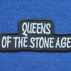 Może przedstawiać: Czarna haftowana naszywka o schodkowym prostokątnym kształcie z białym napisem QUEENS OF THE STONE AGE wykonanym stylizowaną czcionką. Naszywka posiada czarne obszycie i jest przeznaczona do umieszczenia na powierzchniach z tkaniny.