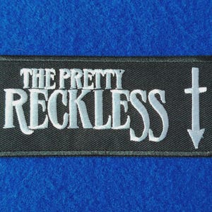 Könnte beinhalten: Ein rechteckiger bestickter Aufnäher mit schwarzem Hintergrund und weißem Rand. Der Text THE PRETTY RECKLESS ist in weißen Großbuchstaben gestickt, gefolgt von einem weißen, nach unten zeigenden Kreuzsymbol auf der rechten Seite des Aufnähers.