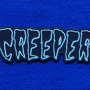 Puede incluir: Un parche bordado que presenta la palabra CREEPER en una fuente estilizada y dentada. Las letras son de color azul claro con un contorno oscuro sobre un fondo negro, creando un diseño de alto contraste para aplicar en telas.