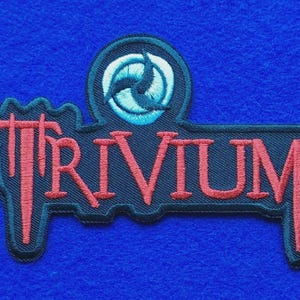 May include: An embroidered patch featuring the word TRIVIUM in red lettering with a black border. Above the text is a circular emblem containing a light blue and white stylised three-pointed symbol on a black background.