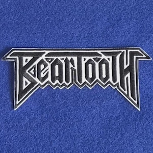 May include: A black and white embroidered patch featuring the word BEARTOOTH in a stylized, angular, heavy metal-inspired font. The design has a sharp, geometric border that follows the contours of the lettering, creating a bold, high-contrast look.