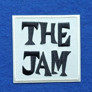May include: A square embroidered patch featuring a white background with a thin white border. The text THE JAM is stitched in bold black capital letters across the centre, with THE on the top line and JAM on the bottom line.