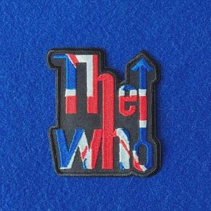 May include: An embroidered fabric patch featuring the text The Who in a stylized font. The letters are filled with a red, white, and blue Union Jack flag pattern against a black background, with an arrow pointing upward from the letter o.