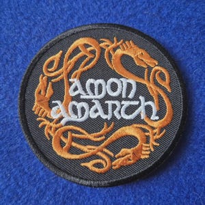 May include: A circular embroidered patch featuring a black background with an orange dragon motif forming a border. The text Amon Amarth is stitched in white across the centre in a stylised font. The patch has a black stitched border edge.