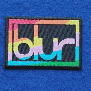 May include: A rectangular embroidered patch featuring the word blur in lowercase letters. The design includes a black background with a multi-coloured border and diagonal stripes in shades of pink, orange, yellow, light purple, and bright blue.