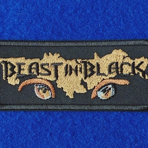 May include: A rectangular black embroidered patch featuring the gold-coloured text BEAST IN BLACK. Below the text are two stylised eyes with orange irises and dark lashes, all rendered in detailed embroidery on a black fabric background.