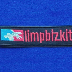 Puede incluir: Un parche rectangular bordado con fondo negro. A la izquierda, un pequeño cuadrado presenta un gráfico de un perro en azul claro y rosa. A la derecha, el texto "limpbizkit" está bordado en letras minúsculas rojas sin remates.