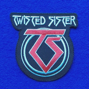 May include: A black embroidered patch featuring the band name Twisted Sister in light blue lettering at the top. Below the text is a red stylised TS logo enclosed within a light blue circular border, all set against a dark fabric background.