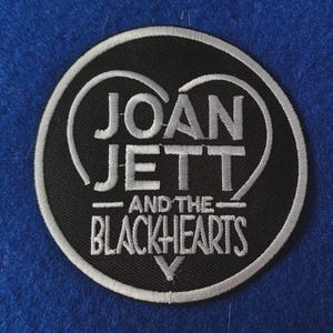Könnte beinhalten: Ein kreisförmiger gestickter Aufnäher mit schwarzem Hintergrund und weißem Rand. Der Text JOAN JETT AND THE BLACKHEARTS ist mittig in weißem Garn gestickt und von einer weißen, gestickten Herzform umrahmt, die den Bandnamen umschließt.