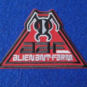 May include: A triangular embroidered patch featuring a red background with a black border. At the top is a stylised black and white ant head icon. Below the icon, the letters AAF are embroidered in black, followed by the text ALIEN ANT FARM in white.