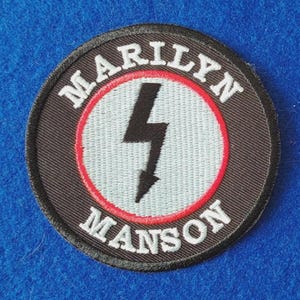 May include: A circular embroidered patch featuring a black border with the white text MARILYN MANSON. The centre is a light grey circle with a thin red ring and a bold black lightning bolt symbol pointing downward in the middle of the design.
